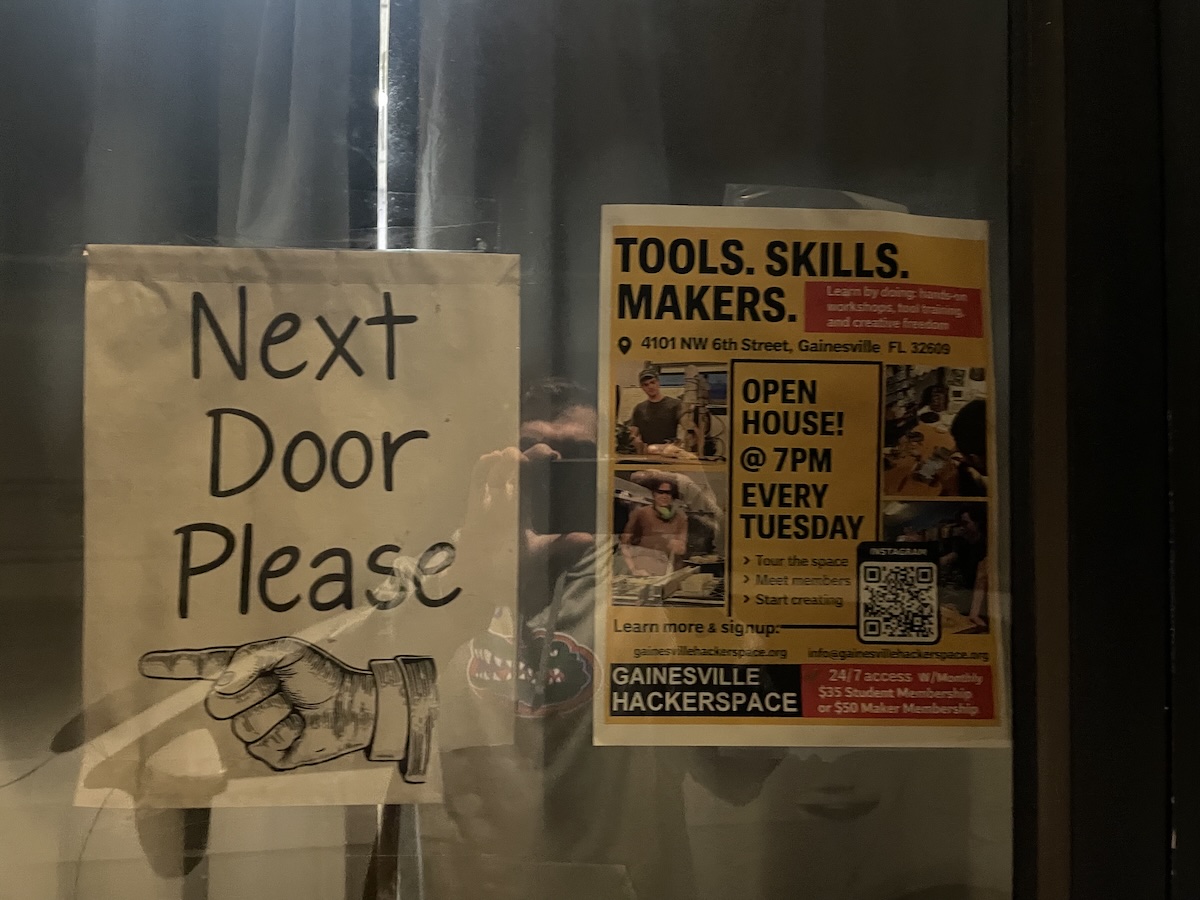 Handwritten sign reading 'Next Door Please' outside a hackerspace, with a faint reflection in the window