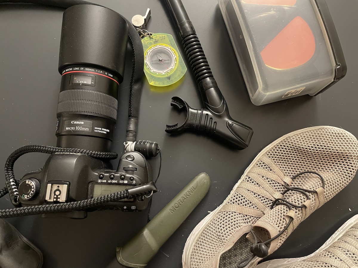 Kayak gear laid out: camera, compass, snorkel mask, knife, and water shoes arranged on a dark surface