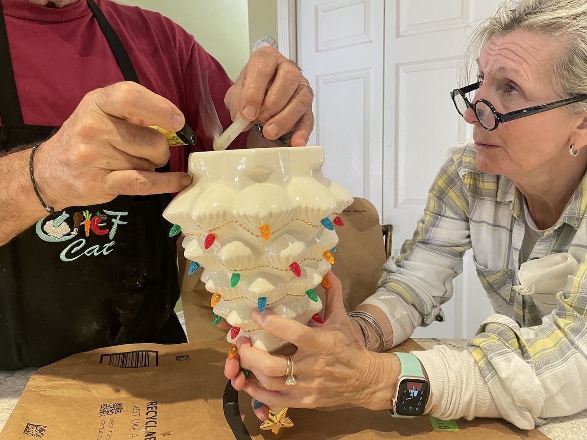 Hands repairing a small porcelain Christmas tree with hot glue at a kitchen table