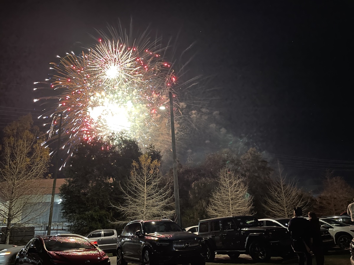 Fireworks over a city park with parked cars and silhouettes in the foreground