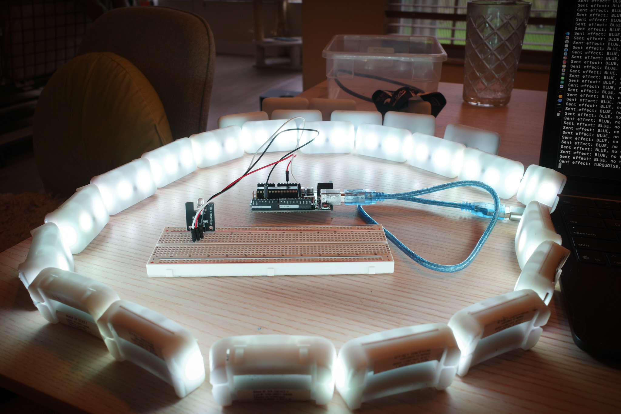 Ring of illuminated PixMob wristband lights surrounding an infrared transmitter connected to a breadboard and microcontroller on a table.